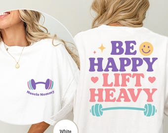 May include: White t-shirt with a purple dumbbell and the text "Muscle Mommy" on the front. The back of the shirt has the text "Be Happy Lift Heavy" in pink, purple, and blue with a yellow smiley face and a barbell.