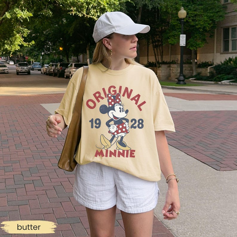 May include: Butter-colored t-shirt featuring a graphic of Minnie Mouse with the text "ORIGINAL 1928 MINNIE". The shirt is paired with white shorts and a white baseball cap. A brown shoulder bag is also visible.