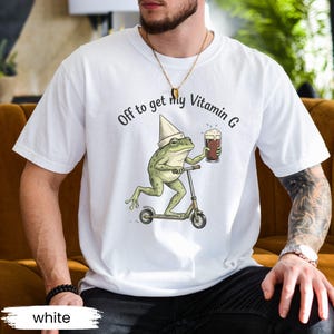 May include: White t-shirt with a whimsical graphic of a frog riding a scooter and holding a beer. The text "Off to get my Vitamin G" is printed above the frog. The shirt is a short-sleeved crew neck.