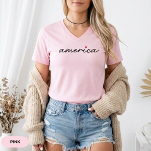 K&ouml;nnte beinhalten: Ein rosa V-Ausschnitt-T-Shirt mit dem Wort "america" in Schreibschrift und einem roten Stern. Das Shirt wird mit Jeansshorts und einer beigen Strickjacke getragen. Das Wort "PINK" befindet sich am unteren Bildrand.