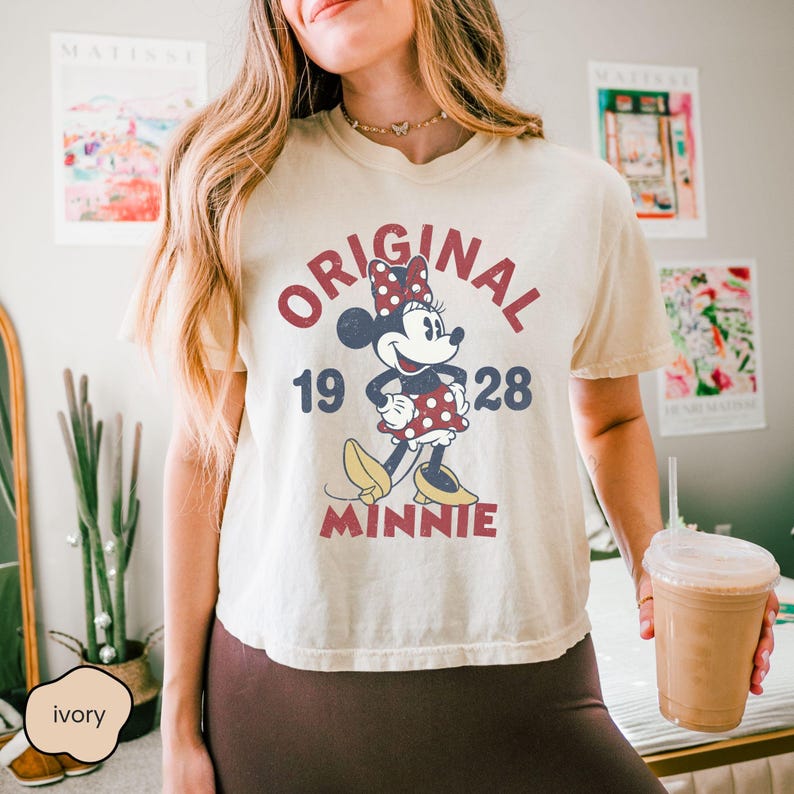 May include: Ivory-colored t-shirt featuring a vintage Minnie Mouse graphic. The design includes the words "ORIGINAL MINNIE 1928" in red and navy blue. Minnie Mouse is depicted in her classic polka-dot dress and yellow shoes.