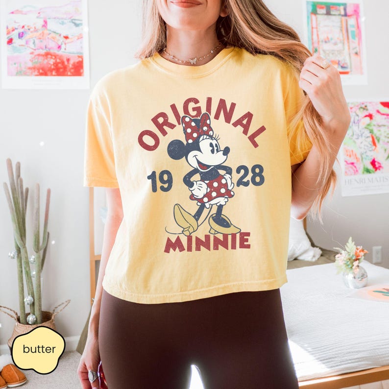 May include: Yellow t-shirt featuring a vintage Minnie Mouse graphic. The design includes the words "ORIGINAL 1928 MINNIE" in a retro font. The shirt is a light yellow color.