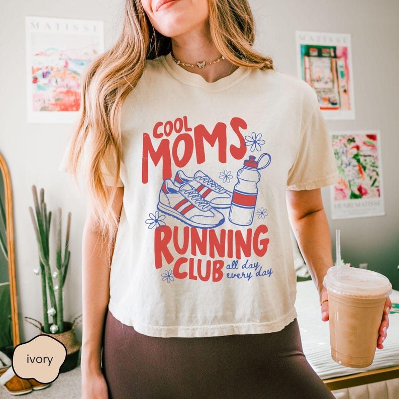 May include: An ivory short-sleeve t-shirt featuring a red and blue graphic print that reads COOL MOMS RUNNING CLUB all day every day, accompanied by illustrations of a pair of athletic sneakers and a water bottle surrounded by small flowers.