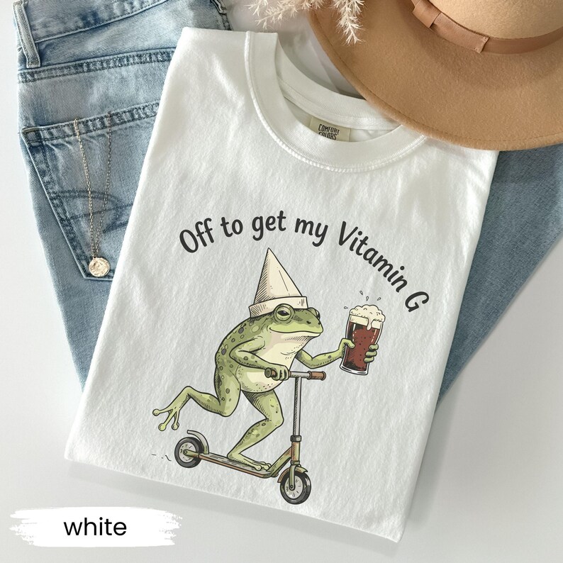 May include: White t-shirt with a graphic of a green frog riding a scooter and holding a beer. The text "Off to get my Vitamin G" is printed above the frog. The shirt is displayed on a pair of blue jeans.