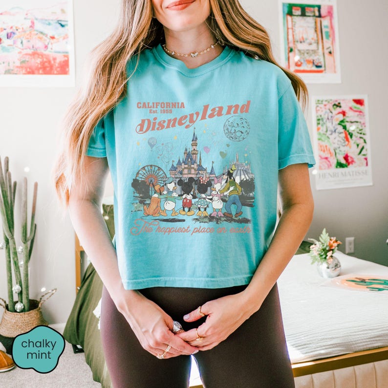 May include: A turquoise t-shirt featuring a Disneyland graphic with the text "California Est. 1955 Disneyland The happiest place on earth." The graphic includes cartoon characters and a castle. The shirt is short-sleeved.