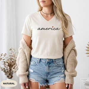 K&ouml;nnte beinhalten: Cremefarbenes V-Ausschnitt-T-Shirt mit dem Wort "america" in schwarzer Schrift und einem roten Stern. Das Shirt wird mit Jeansshorts und einer beigen Strickjacke kombiniert. Das Wort "NATURAL" steht unten links.