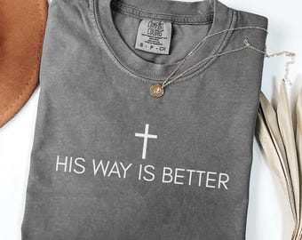 May include: A gray Comfort Colors t-shirt with the text "HIS WAY IS BETTER" and a cross graphic. A gold-colored necklace with a pendant rests on the shirt. A brown hat and dried leaves are in the background.