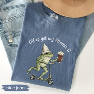 May include: A blue Comfort Colors t-shirt with the phrase "Off to get my Vitamin G" and a cartoon frog riding a scooter while holding a beer. The frog wears a white hat. The shirt is displayed on a pair of blue jeans.
