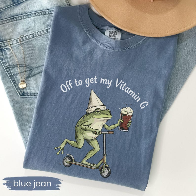 May include: A blue Comfort Colors t-shirt with the phrase "Off to get my Vitamin G" and a cartoon frog riding a scooter while holding a beer. The frog wears a white hat. The shirt is displayed on a pair of blue jeans.
