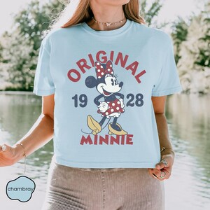 May include: Light blue t-shirt featuring a vintage-style Minnie Mouse graphic. The design includes the words "ORIGINAL 1928 MINNIE" in red, with Minnie Mouse wearing a polka-dot dress and bow. The shirt has short sleeves and a crew neck.