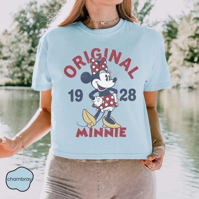 May include: Light blue t-shirt featuring a vintage-style Minnie Mouse graphic. The design includes the words "ORIGINAL 1928 MINNIE" in red, with Minnie Mouse wearing a polka-dot dress and bow. The shirt has short sleeves and a crew neck.