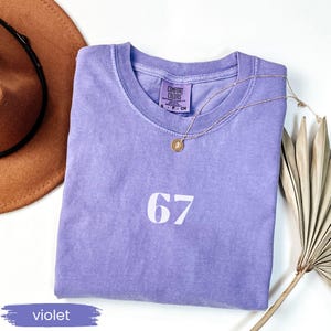 May include: A violet Comfort Colors t-shirt with the number 67 printed in white. A gold necklace with a pendant rests on the shirt. A brown hat and dried palm leaf are in the background.