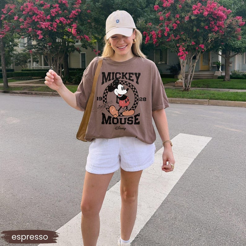 May include: Espresso-colored t-shirt featuring a vintage Mickey Mouse graphic. The shirt has a crew neck and short sleeves. The image also shows white shorts and a beige baseball cap. The overall style is casual and comfortable.