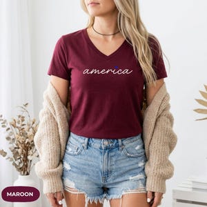 K&ouml;nnte beinhalten: Burgunderrotes V-Ausschnitt-T-Shirt mit dem Wort &bdquo;america&ldquo; in Schreibschrift. Ein kleiner blauer Punkt ersetzt den Punkt &uuml;ber dem &bdquo;i&ldquo;. Das Model tr&auml;gt Jeansshorts und eine beige Strickjacke.