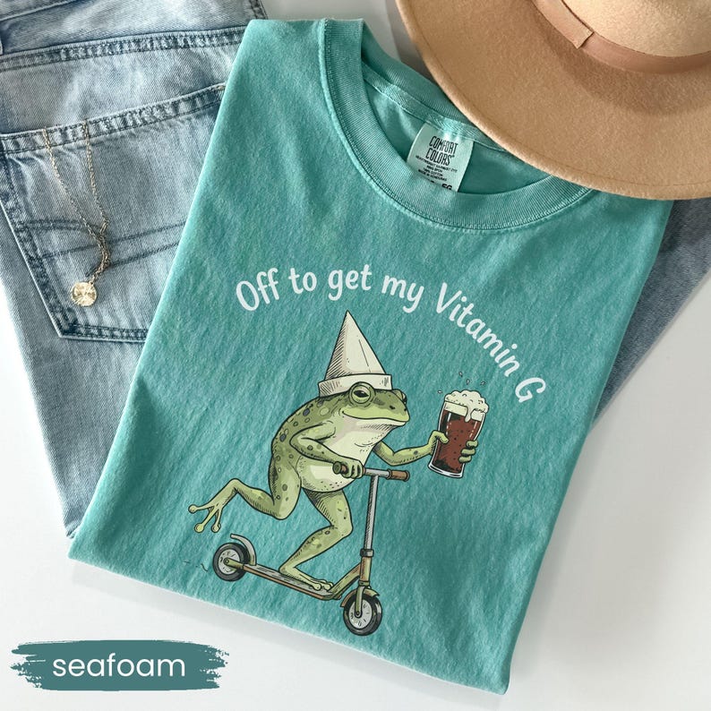May include: Seafoam green t-shirt with the phrase "Off to get my Vitamin G." The shirt features a cartoon frog riding a scooter and holding a beer. The shirt is paired with blue jeans and a tan hat.