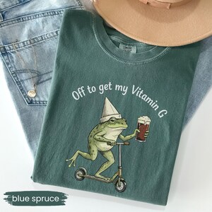 May include: Blue spruce t-shirt with a frog graphic riding a scooter and holding a beer. The text "Off to get my Vitamin G" is printed above the frog. The shirt is paired with light blue jeans and a tan hat.