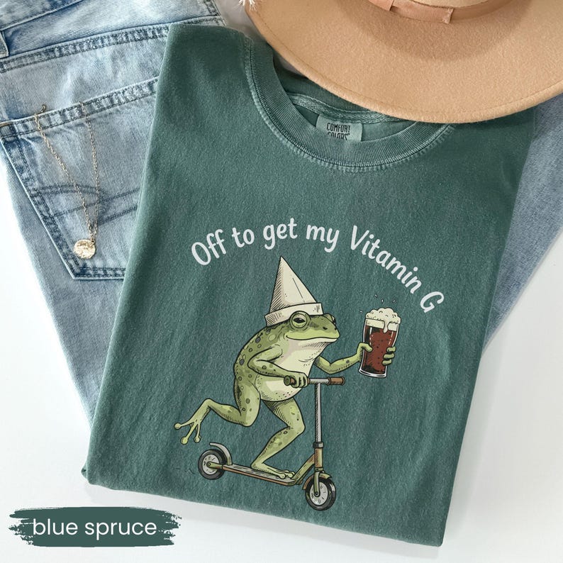 May include: Blue spruce t-shirt with a frog graphic riding a scooter and holding a beer. The text "Off to get my Vitamin G" is printed above the frog. The shirt is paired with light blue jeans and a tan hat.