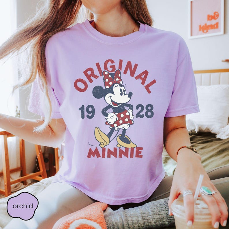 May include: A light purple t-shirt featuring a vintage-style graphic of Minnie Mouse. The design includes the word "ORIGINAL" above the character, the year "1928", and the name "MINNIE" below. The shirt has short sleeves.