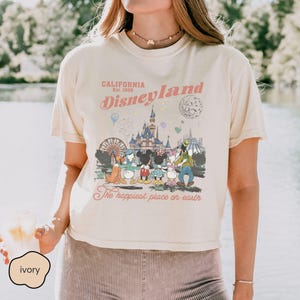 May include: Ivory-colored t-shirt featuring a Disneyland graphic. The design includes the text "California Est. 1955 Disneyland" in pink and a colorful illustration of Disney characters and park attractions. The phrase "The happiest place on earth" is also included.
