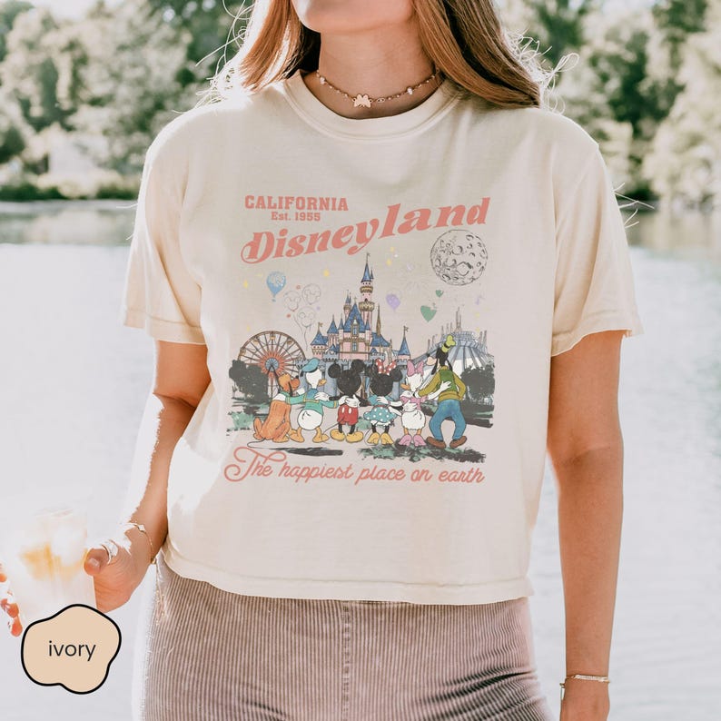 May include: Ivory-colored t-shirt featuring a Disneyland graphic. The design includes the text "California Est. 1955 Disneyland" in pink and a colorful illustration of Disney characters and park attractions. The phrase "The happiest place on earth" is also included.