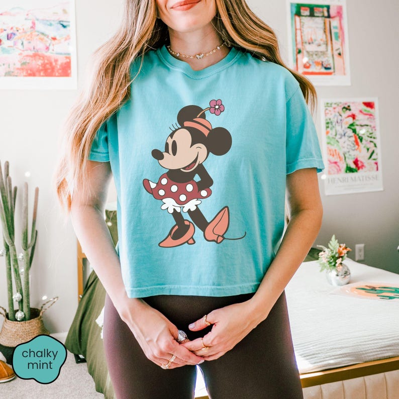 Op de afbeelding: Een lichtblauw T-shirt met een Minnie Mouse-afbeelding. Het stripfiguur draagt een rode polkadot rok, roze schoenen en een bijpassende hoed met een bloem. Het shirt heeft een ronde hals en korte mouwen.