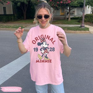 May include: A light pink t-shirt featuring a graphic of Minnie Mouse with the text "ORIGINAL MINNIE 1928". The shirt has short sleeves and a crew neck. The word "blossom" is in a pink rectangle at the bottom left.