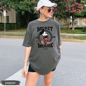 May include: Pepper-colored, oversized t-shirt featuring a graphic of Mickey Mouse. The design includes the text "Mickey Mouse" and the year "1928" in a circular frame. The shirt is a casual style, suitable for everyday wear.