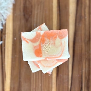 May include: Three bars of handmade soap with a swirl pattern in shades of white and orange. The top bar has a small, gold-colored letter "R" stamped on it.