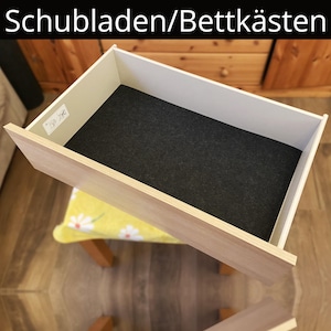 Könnte beinhalten: Eine offene Holzschublade mit einer dunkelgrauen Filzverkleidung. Die Schublade ist oben mit weißem Text "Schubladen/Bettkästen" beschriftet. Die Schublade ist angewinkelt und zeigt ihr Inneres und ihre Konstruktion.