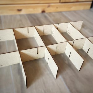 Peut inclure: Un organisateur en bois clair, composé de plusieurs compartiments. Il est fabriqué à partir de fines pièces rectangulaires en bois, avec des fentes et des languettes pour l'assemblage. Le texte "T PRT-007" est imprimé sur le côté.