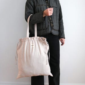 May include: A large, beige tote bag with drawstring closure and long handles. The bag is held by a person wearing a dark grey jacket and black trousers. The bag has a small label on the front.