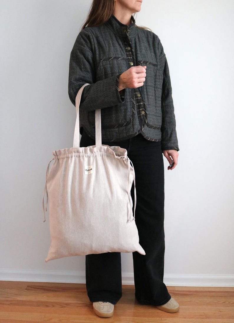 May include: A large, beige tote bag with drawstring closure and long handles. The bag is held by a person wearing a dark grey jacket and black trousers. The bag has a small label on the front.