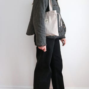 May include: A light beige tote bag with a drawstring closure and a small label. The bag is carried over the shoulder. The person is wearing a dark grey quilted jacket and black trousers. The background is a plain white wall and wooden floor.