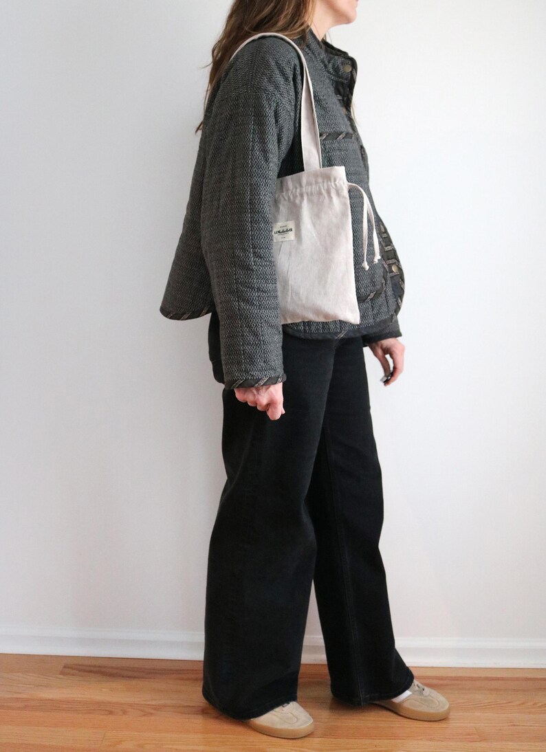 May include: A light beige tote bag with a drawstring closure and a small label. The bag is carried over the shoulder. The person is wearing a dark grey quilted jacket and black trousers. The background is a plain white wall and wooden floor.
