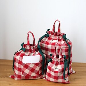 May include: Three red and white gingham drawstring bags of different sizes. Each bag features a green drawstring and handle. The bags have a white label on the front. The bags are on a wooden surface.