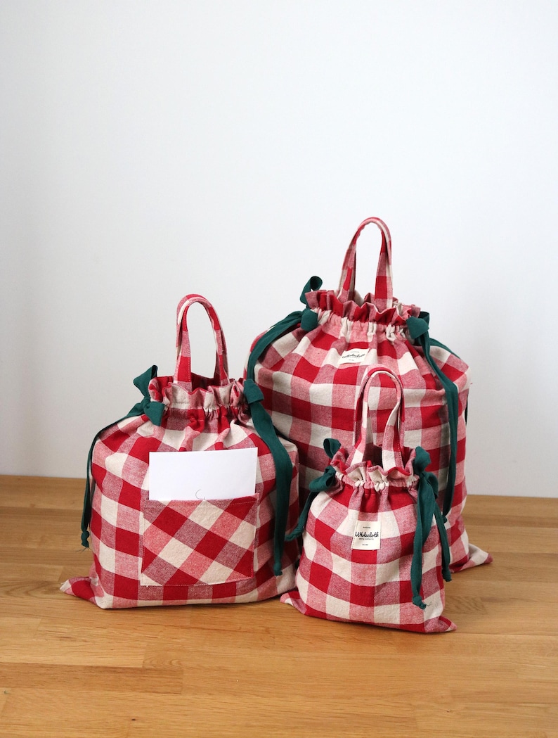 May include: Three red and white gingham drawstring bags of different sizes. Each bag features a green drawstring and handle. The bags have a white label on the front. The bags are on a wooden surface.