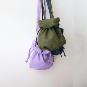 Può includere: Una collezione di piccole borse con coulisse in vari colori, tra cui lavanda, verde oliva e blu denim. Ogni borsa è sospesa da un cinturino in tessuto abbinato, che mostra un design semplice e minimalista. Le borse sono realizzate in un tessuto morbido e strutturato.