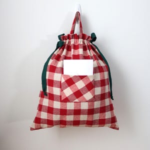 May include: A red and white checked drawstring bag with a green trim and handle, hanging on a white wall. The bag has a pocket with a white card inside. The bag is made of fabric.