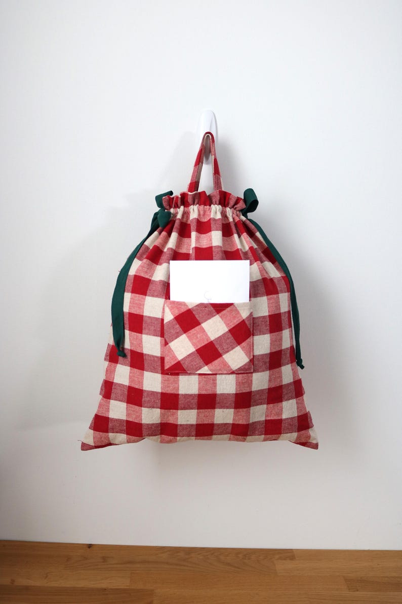 May include: A red and white checked drawstring bag with a green trim and handle, hanging on a white wall. The bag has a pocket with a white card inside. The bag is made of fabric.