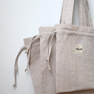May include: Beige linen tote bag with matching drawstring pouches. The tote bag has long handles and a rectangular pocket with a small label. The bags are made of a natural, textured fabric.