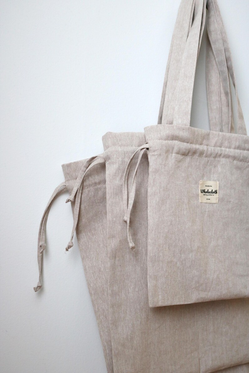 May include: Beige linen tote bag with matching drawstring pouches. The tote bag has long handles and a rectangular pocket with a small label. The bags are made of a natural, textured fabric.