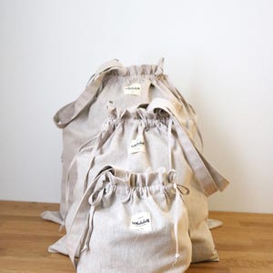 May include: Three beige drawstring bags of different sizes are displayed. Each bag has a matching drawstring closure and a small white label. The bags are made of a light-coloured fabric and are on a wooden surface.