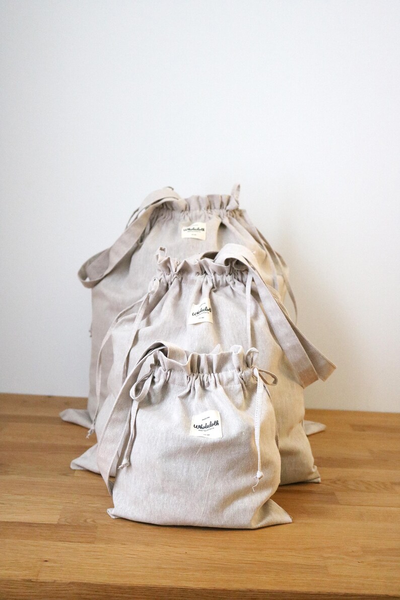May include: Three beige drawstring bags of different sizes are displayed. Each bag has a matching drawstring closure and a small white label. The bags are made of a light-coloured fabric and are on a wooden surface.