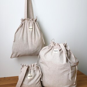 May include: Three beige drawstring bags of different sizes are shown against a white wall and a wooden surface. The bags have a simple design with a small label and a gathered top. One bag is hanging from a hook.