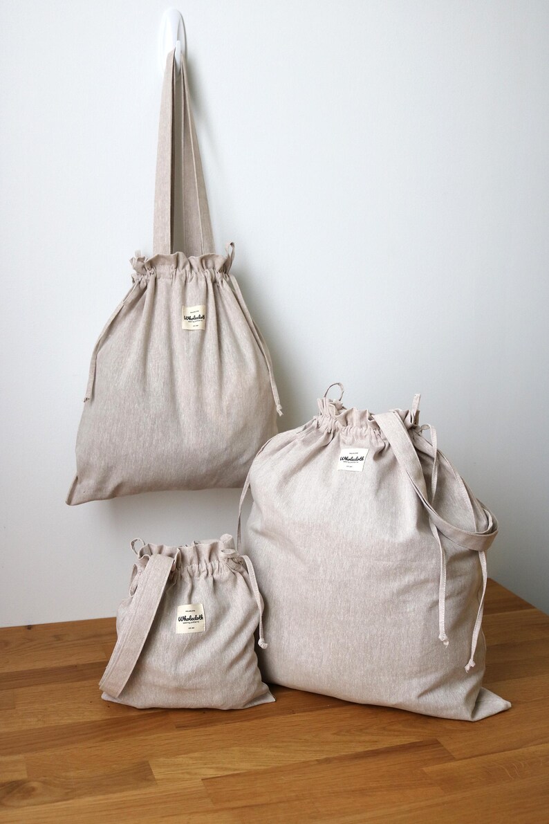 May include: Three beige drawstring bags of different sizes are shown against a white wall and a wooden surface. The bags have a simple design with a small label and a gathered top. One bag is hanging from a hook.
