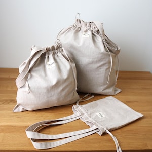 May include: A set of beige linen drawstring bags and a tote bag. The bags are in different sizes, each with a small label. They are made from a natural-coloured fabric and are displayed on a wooden surface.
