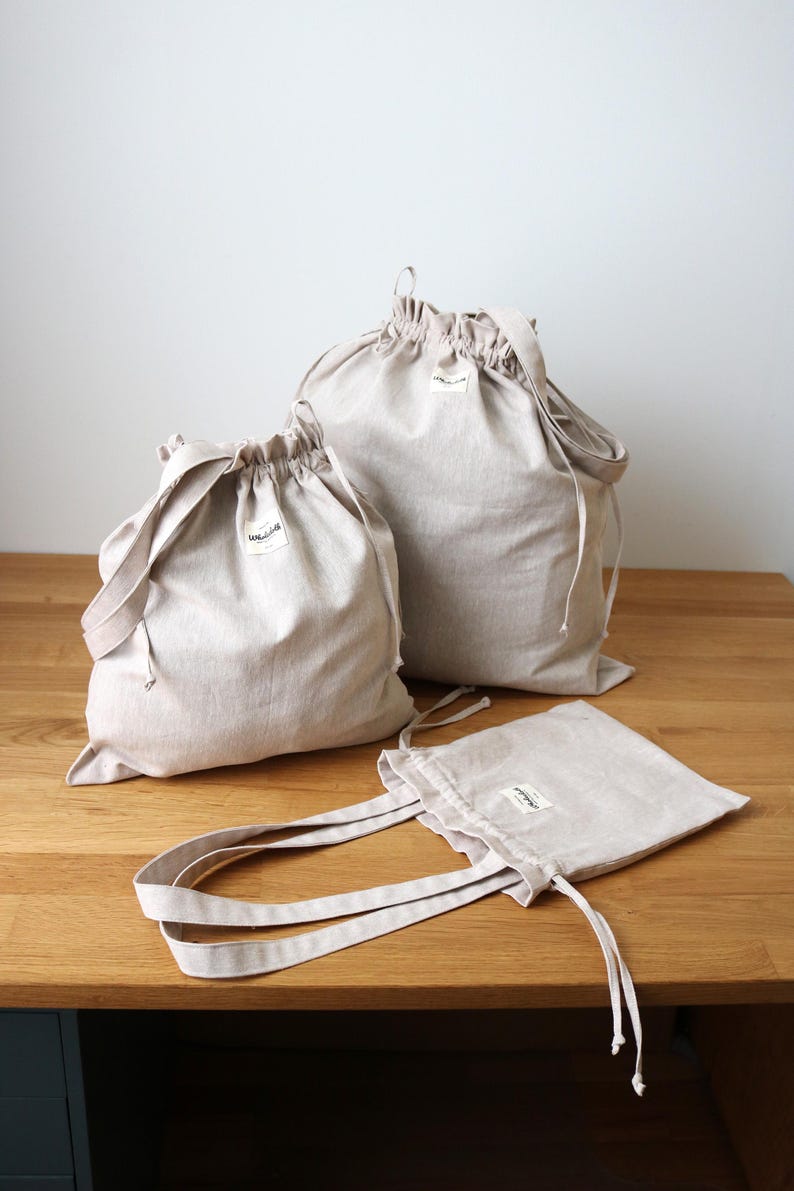May include: A set of beige linen drawstring bags and a tote bag. The bags are in different sizes, each with a small label. They are made from a natural-coloured fabric and are displayed on a wooden surface.