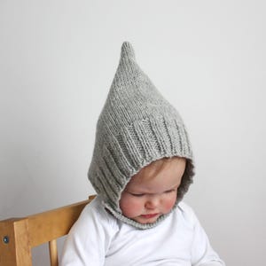 May include: A gray knitted baby hat with a pointed top. The hat is pulled down over the baby's head, covering their ears and forehead.