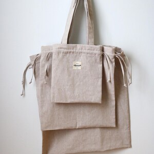 May include: A collection of beige linen tote bags and drawstring pouches suspended from a white wall. The tote bag features a long handle and a small label. The pouches have drawstrings. The bags are made of a natural, textured fabric.