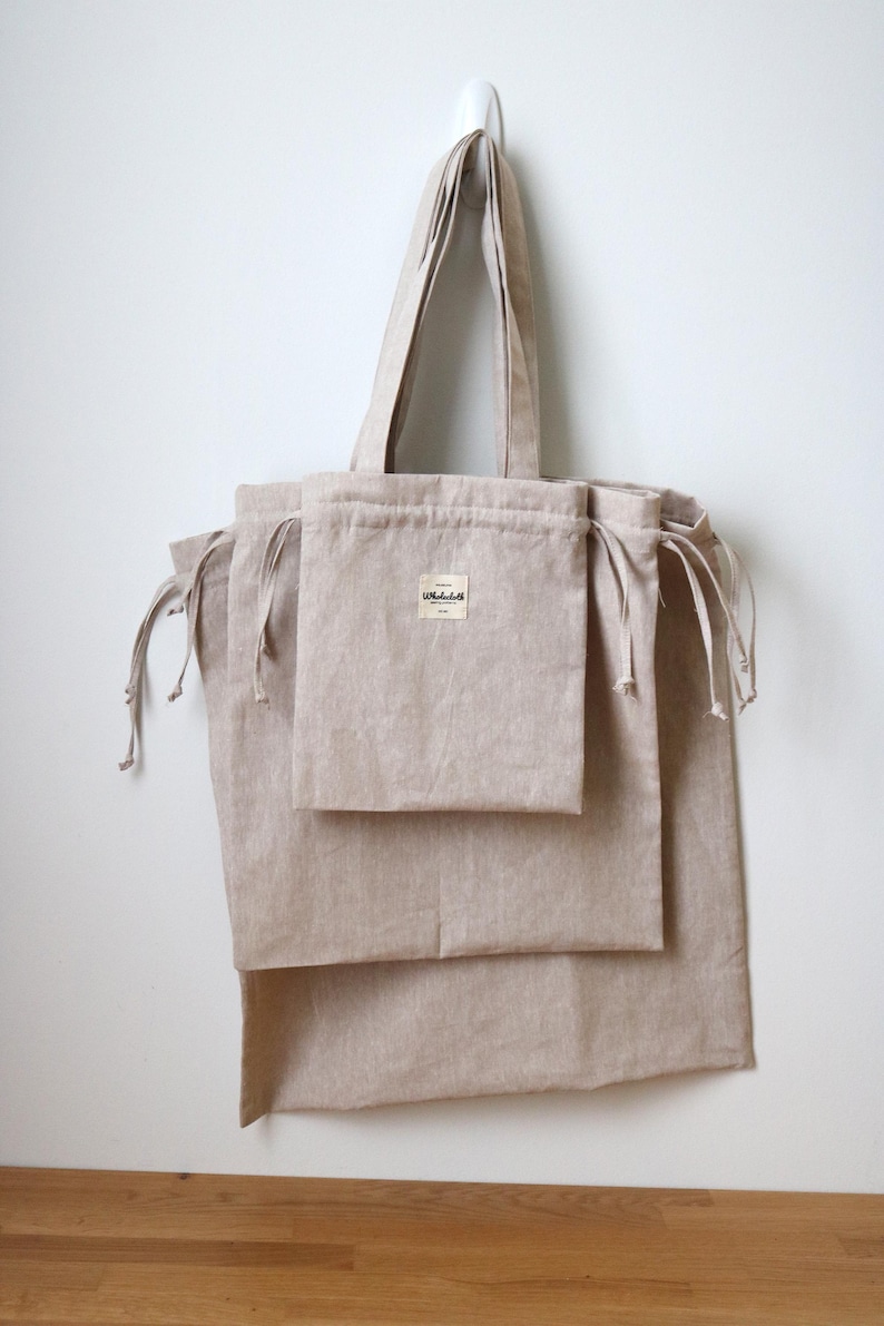 May include: A collection of beige linen tote bags and drawstring pouches suspended from a white wall. The tote bag features a long handle and a small label. The pouches have drawstrings. The bags are made of a natural, textured fabric.